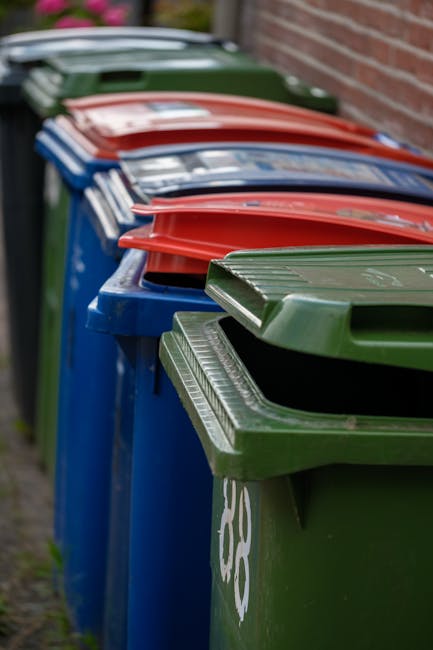 A row of large wheelie bins positioned outdoors along a pavement, with the closest bin in the foreground being green, featuring a textured, ribbed lid and a smooth finish, and the nearby bin showing a