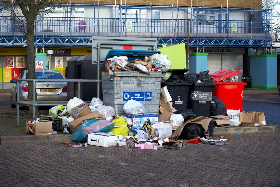 A large collection of mixed waste including cardboard boxes, paper bags, plastic bags, black rubbish bags, and loose paper, all piled on a paved sidewalk next to a metal railing in front of commercial