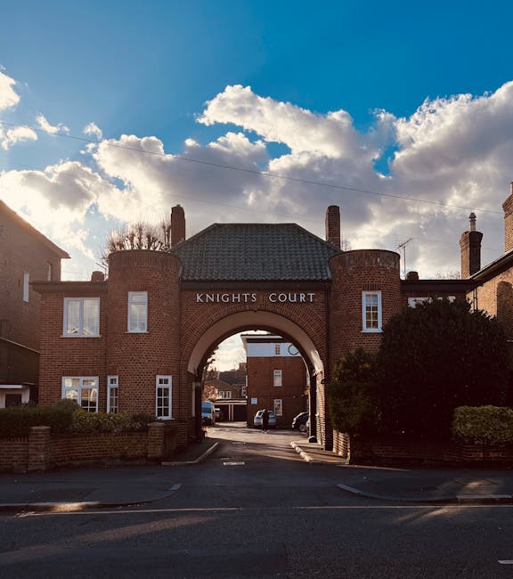 A large brick archway with the inscription 'KNIGHTS COURT' at the top, situated within a residential area, with surrounding red-brick buildings and green bushes on both sides. The archway frames a vie
